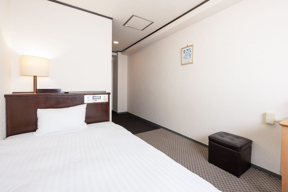 A hotel room with a bed, a nightstand, a lamp, a small black trash bin, and a picture on the wall.