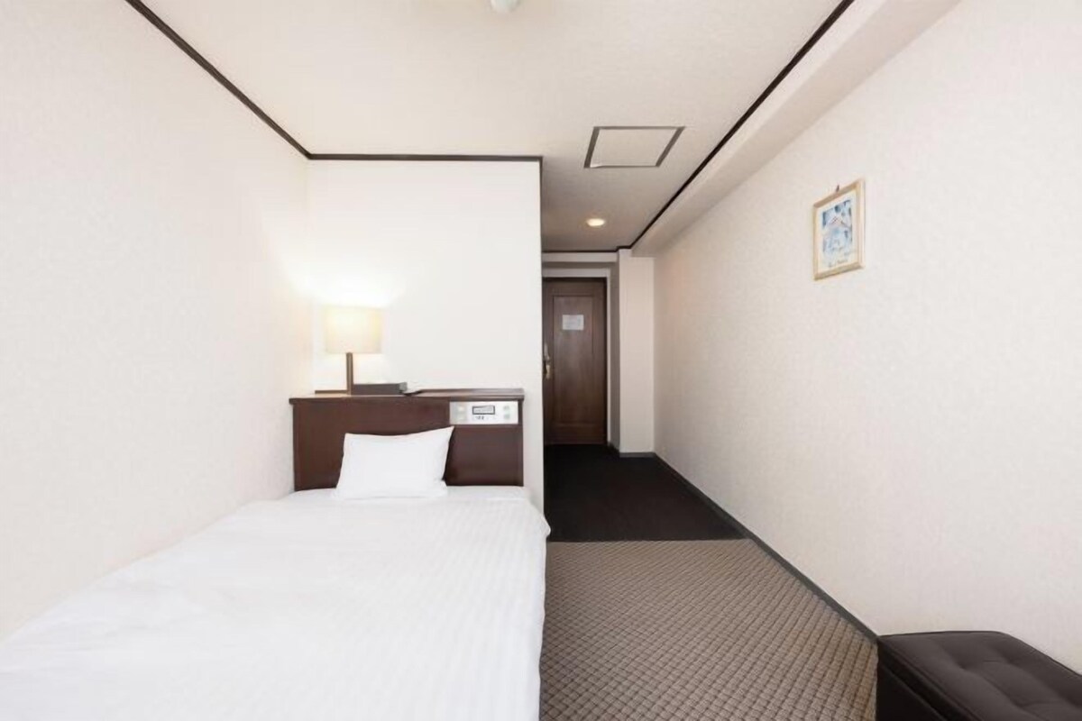 A hotel room with a bed, bedside table, lamp, and a small desk with a chair.