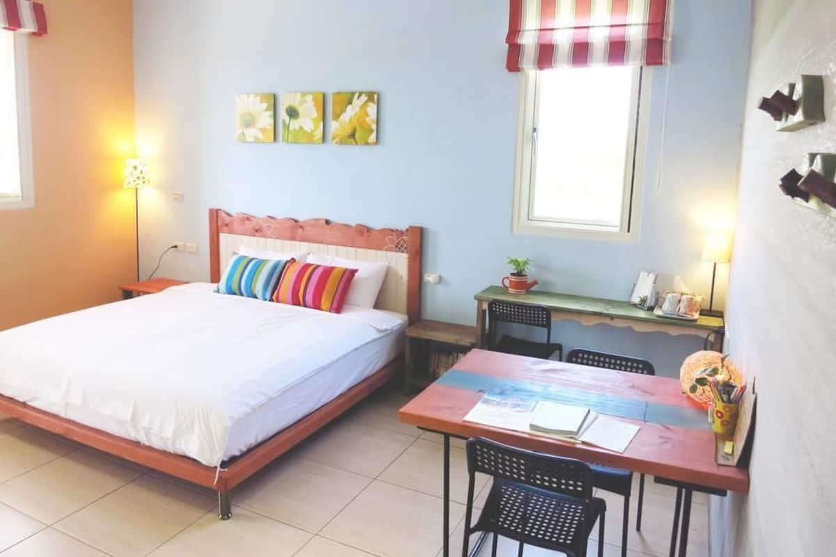 A bedroom with a bed, a desk, and chairs. There are two windows with curtains, a wall clock, and a small table with a plant.