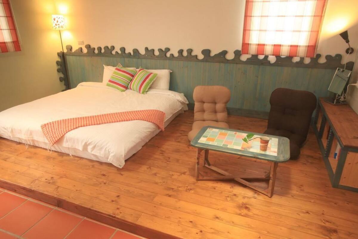 A cozy bedroom with a wooden floor, a bed with striped pillows, a small table with a cup, and a chair.