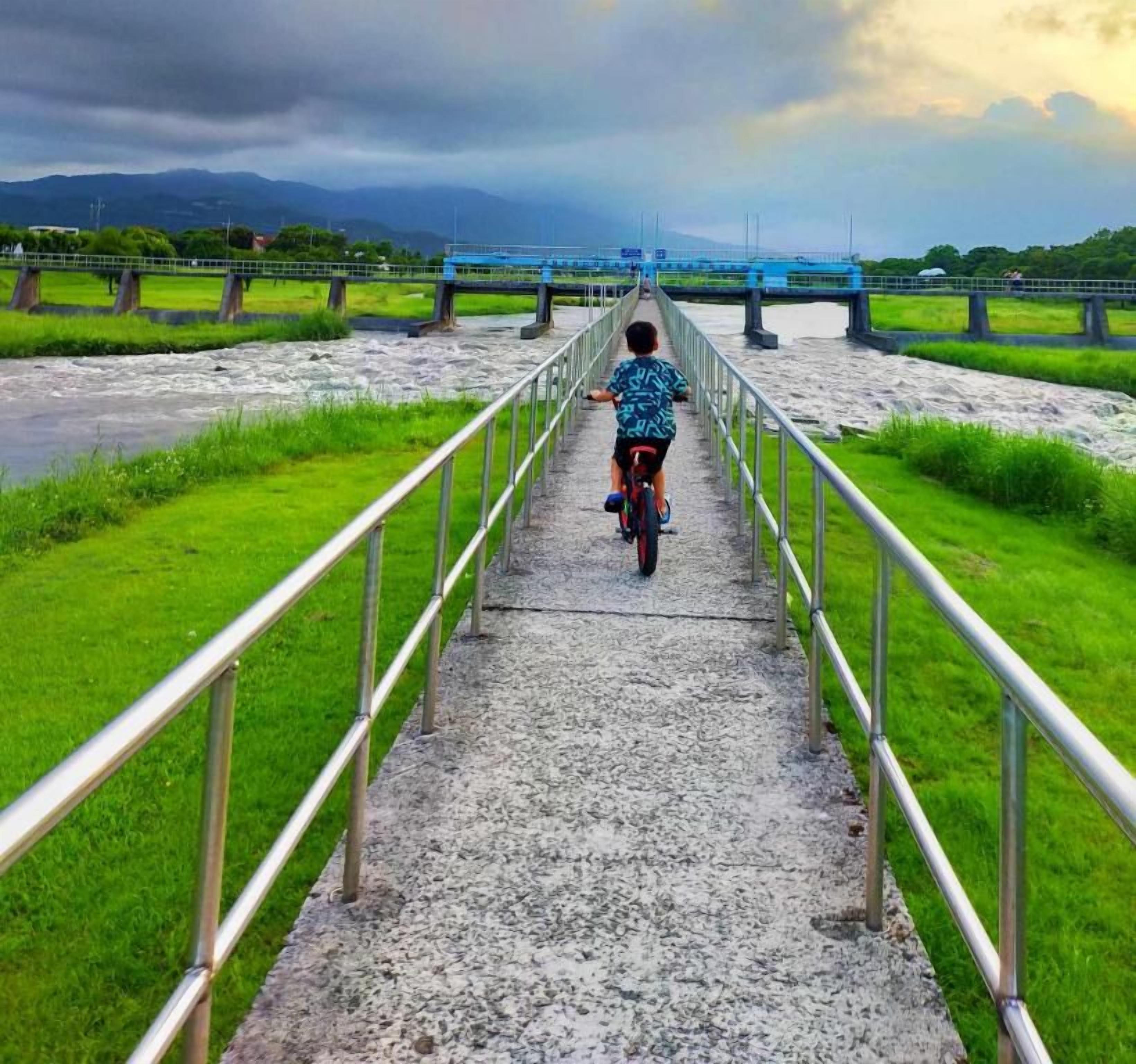 A person riding a bicycle on a concrete pathway with metal railings, leading towards a bridge over a river.