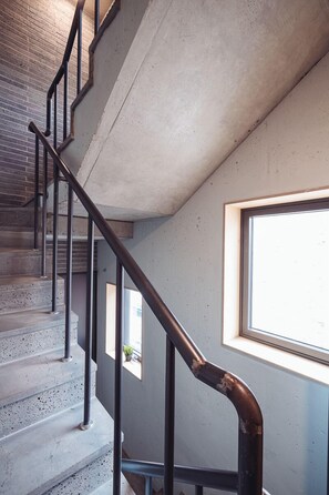 Uma escadaria com degraus de concreto e corrimãos de metal preto, que leva a um quarto com janela e vista para o exterior.