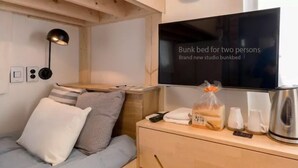 Un lit superposé avec un téléviseur fixé au-dessus, une horloge au mur et une table de chevet avec une bouilloire et un sac de pain.