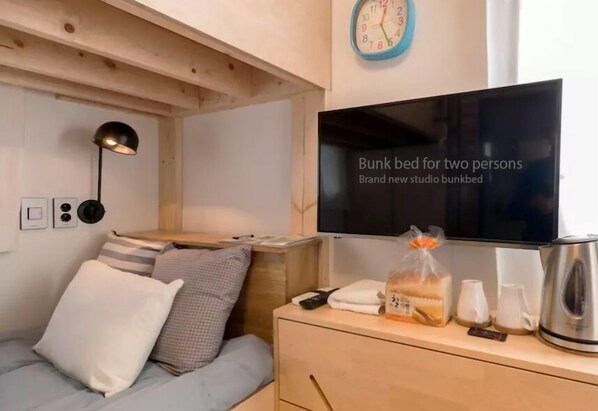 A bunk bed with a television above it, a clock on the wall, and a wooden dresser with a kettle and a bag of bread.