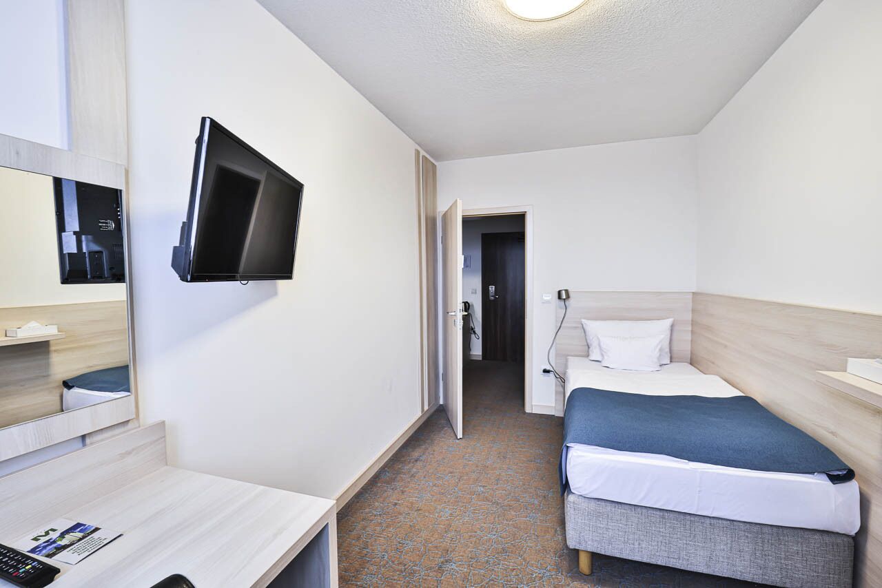 A hotel room with a bed, bedside table, a flat-screen TV mounted on the wall, and a door leading to another room.