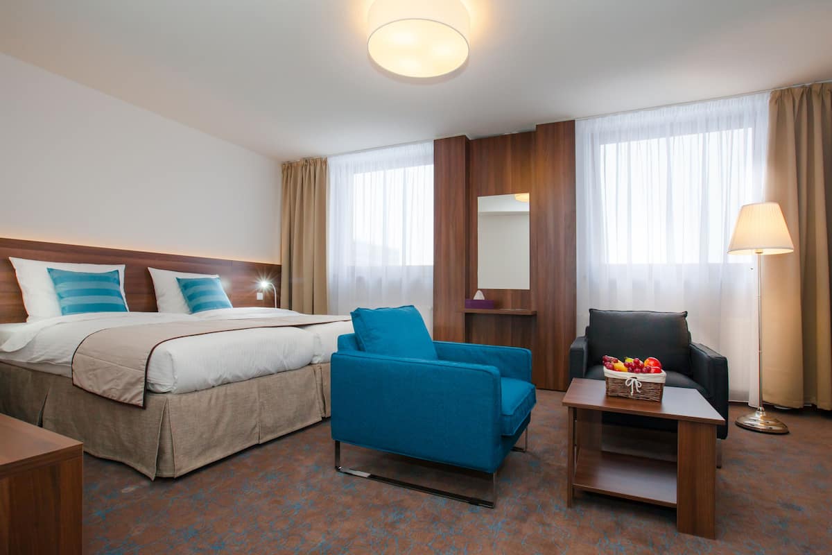 A hotel room with a bed, a blue armchair, a small table with a fruit basket, and a lamp.