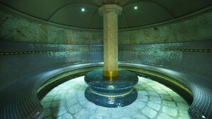Una piscina grande y circular, con mosaicos, y una columna central, rodeada por un suelo de piedra y un techo alto y abovedado.