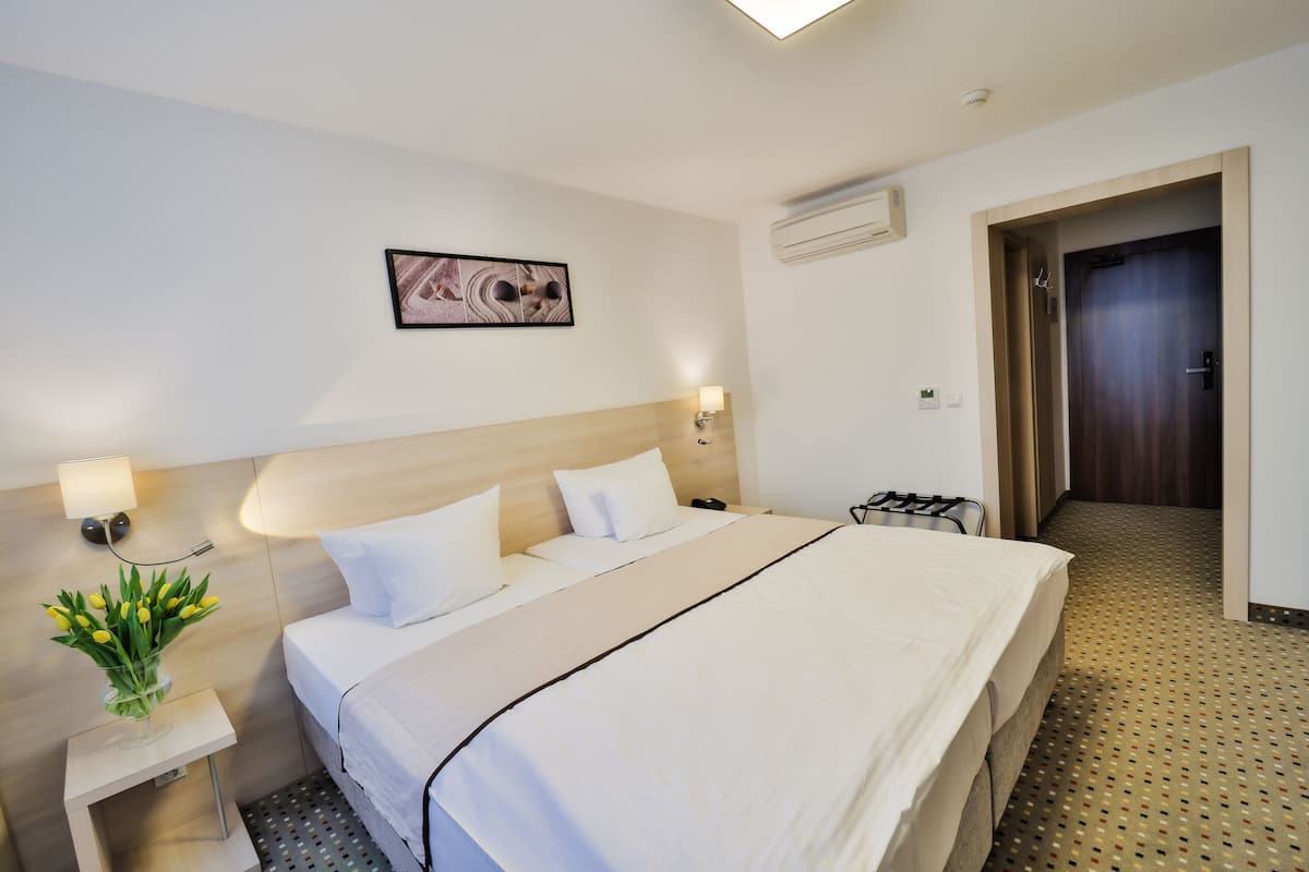 A hotel room with a bed, bedside lamps, a wall-mounted air conditioner, a door, and a small table with a vase of flowers.