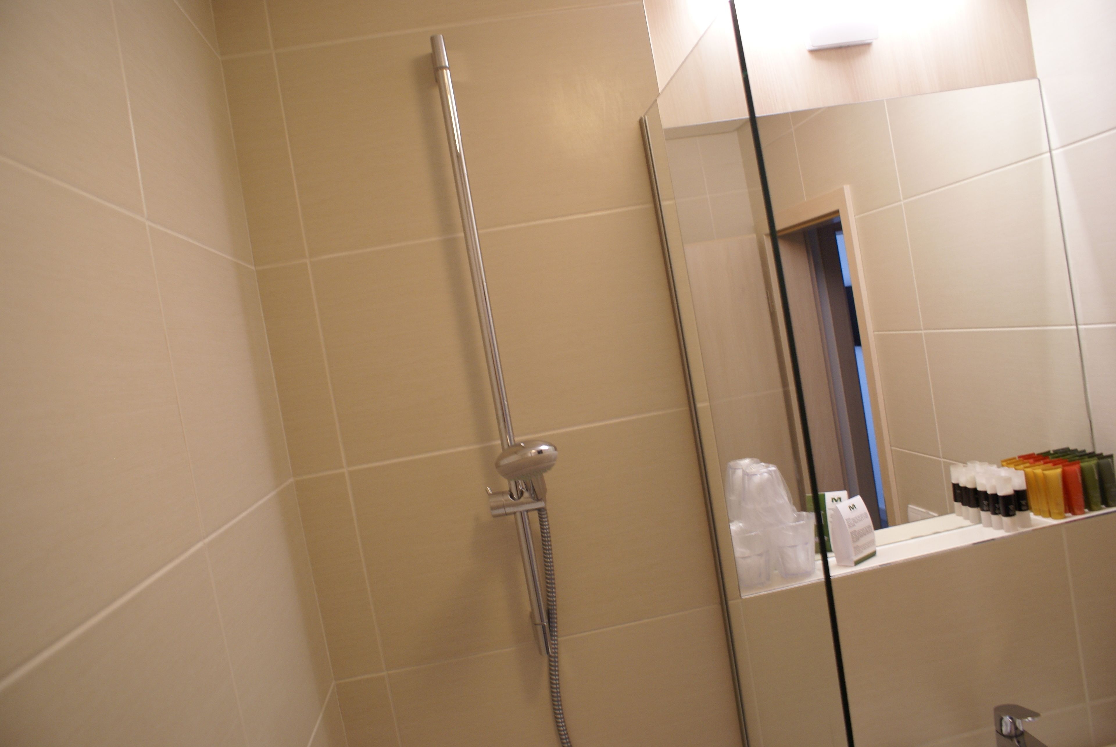 A bathroom with beige tiles, a shower with a handheld showerhead, and a mirror reflecting a door and a shelf with toiletries.