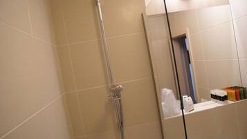 A bathroom with beige tiles, a shower with a handheld showerhead, and a mirror reflecting a door and a shelf with toiletries.