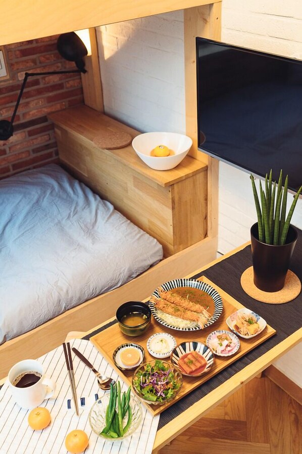 A compact room with a bed, a small table with a tray of food, a TV, and a potted plant.