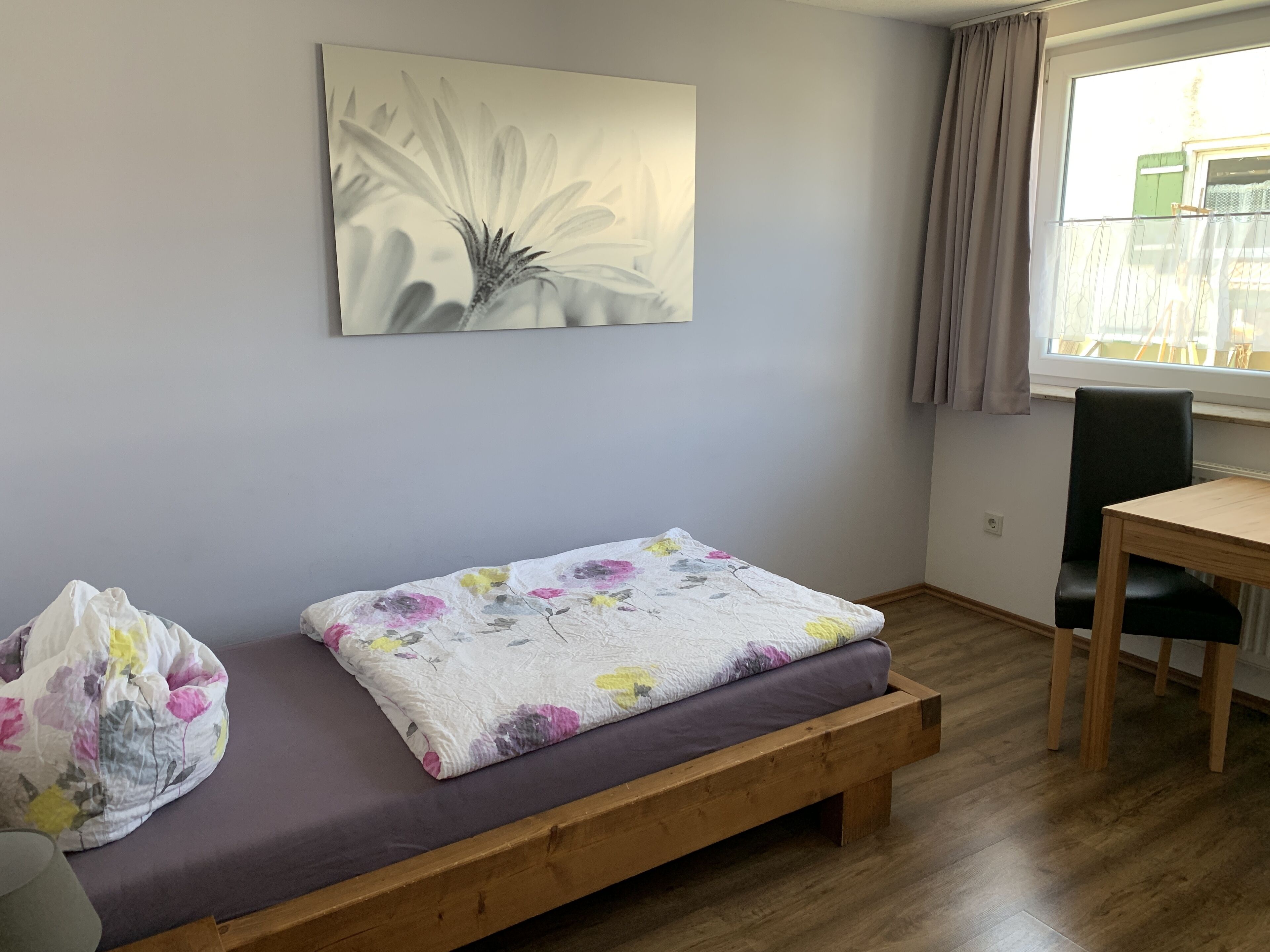 Una habitación con una cama de banco de madera, un cojín estampado con motivos florales, una silla verde y una ventana con cortinas.