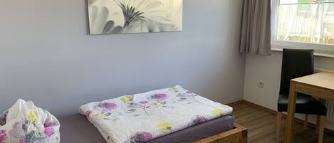 Una habitación con una cama de banco de madera, un cojín estampado con motivos florales, una silla verde y una ventana con cortinas.
