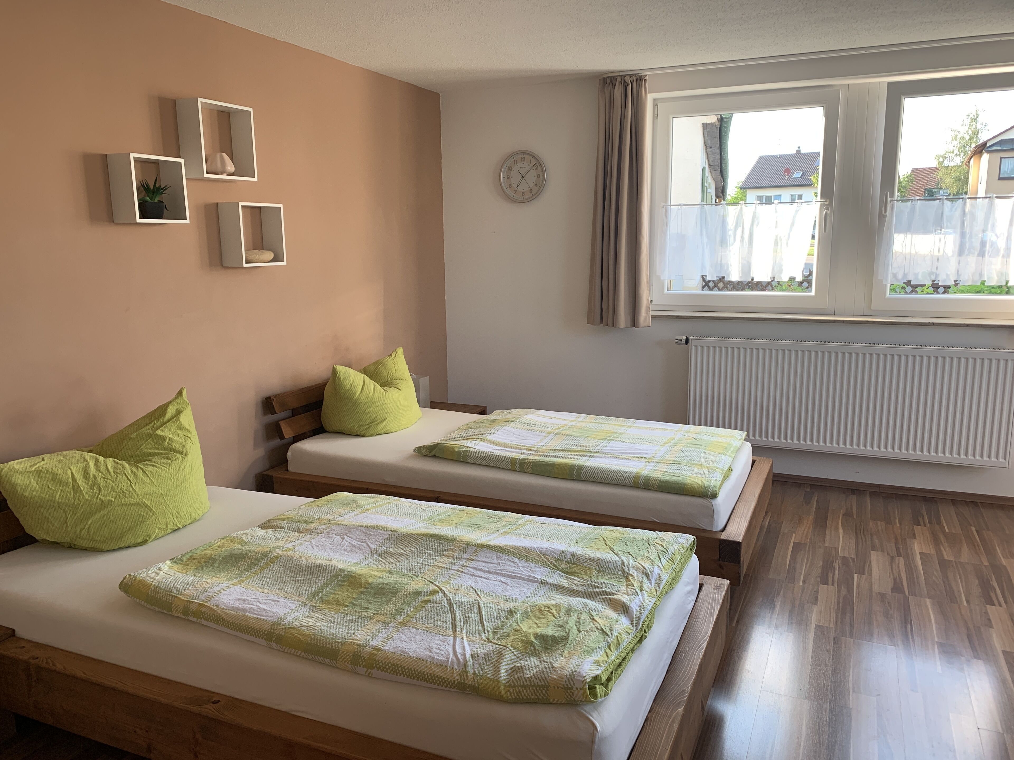A room with two beds, each with a green pillow and a patterned blanket. There are wall-mounted shelves with decorative items, a clock, and a window with curtains.