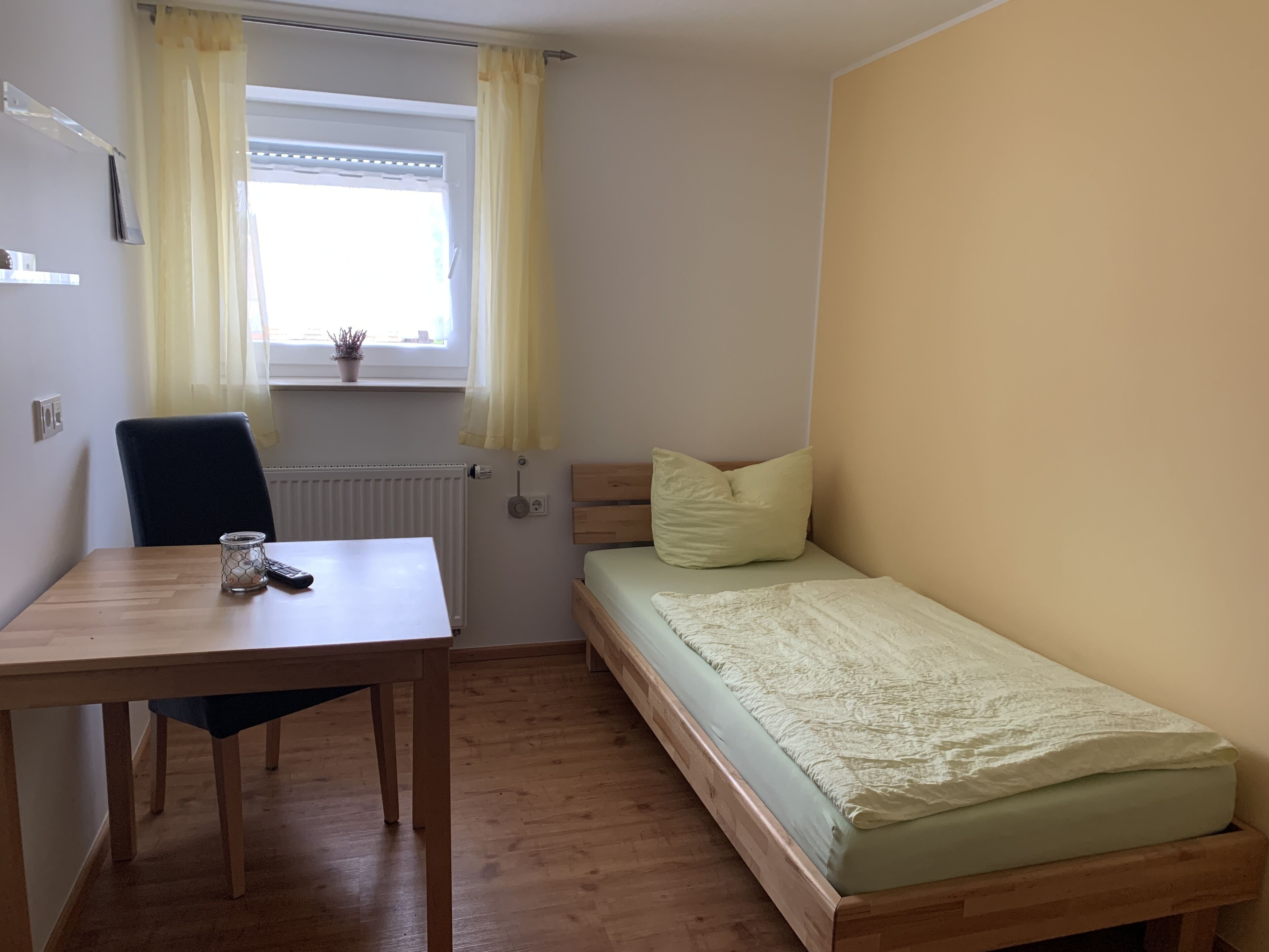 A small room with a wooden table, a single bed, a chair, a window with curtains, and a radiator.