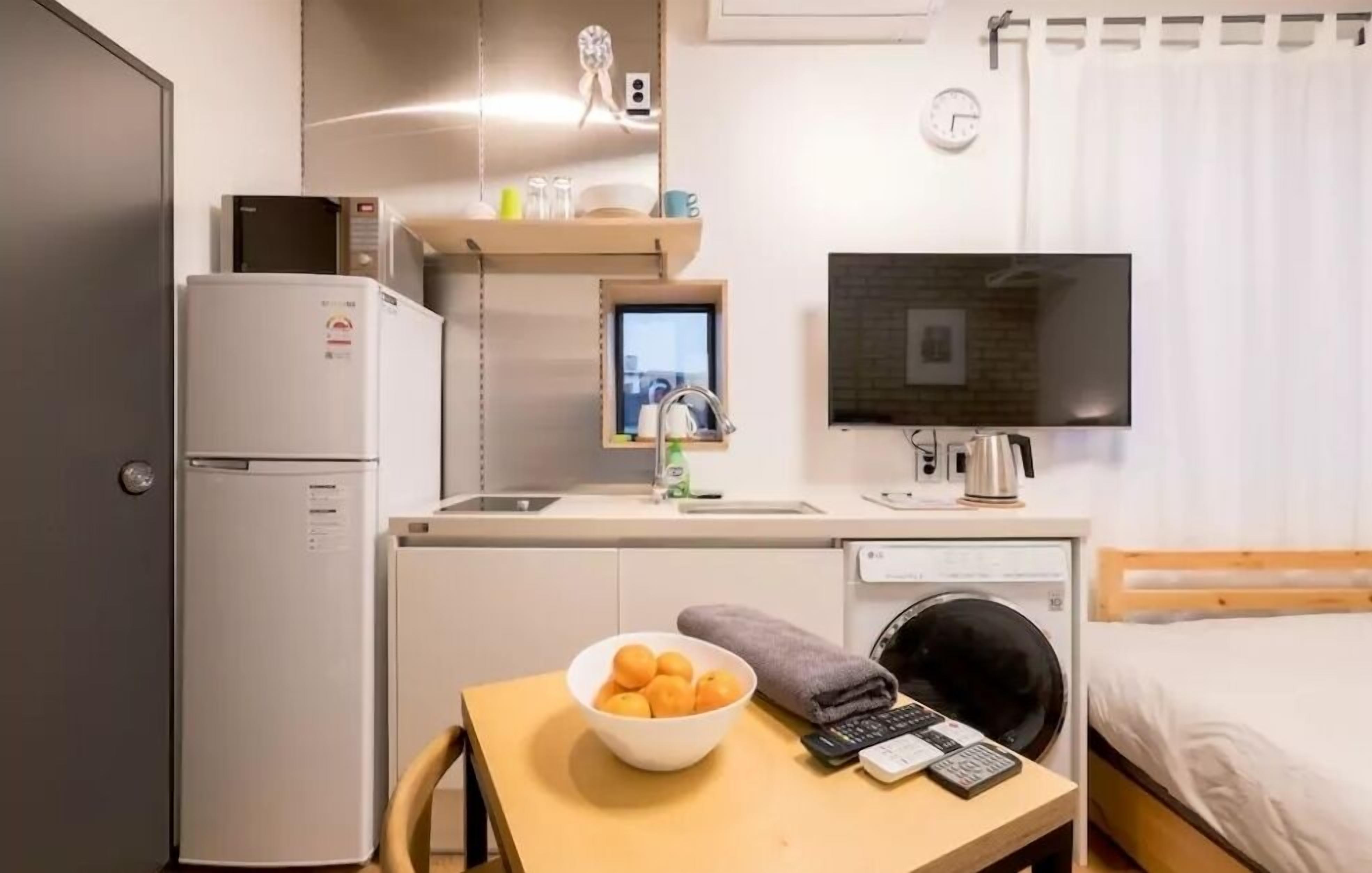 A compact hotel room with a kitchenette, a single bed, a TV, and a small table with a bowl of fruit.