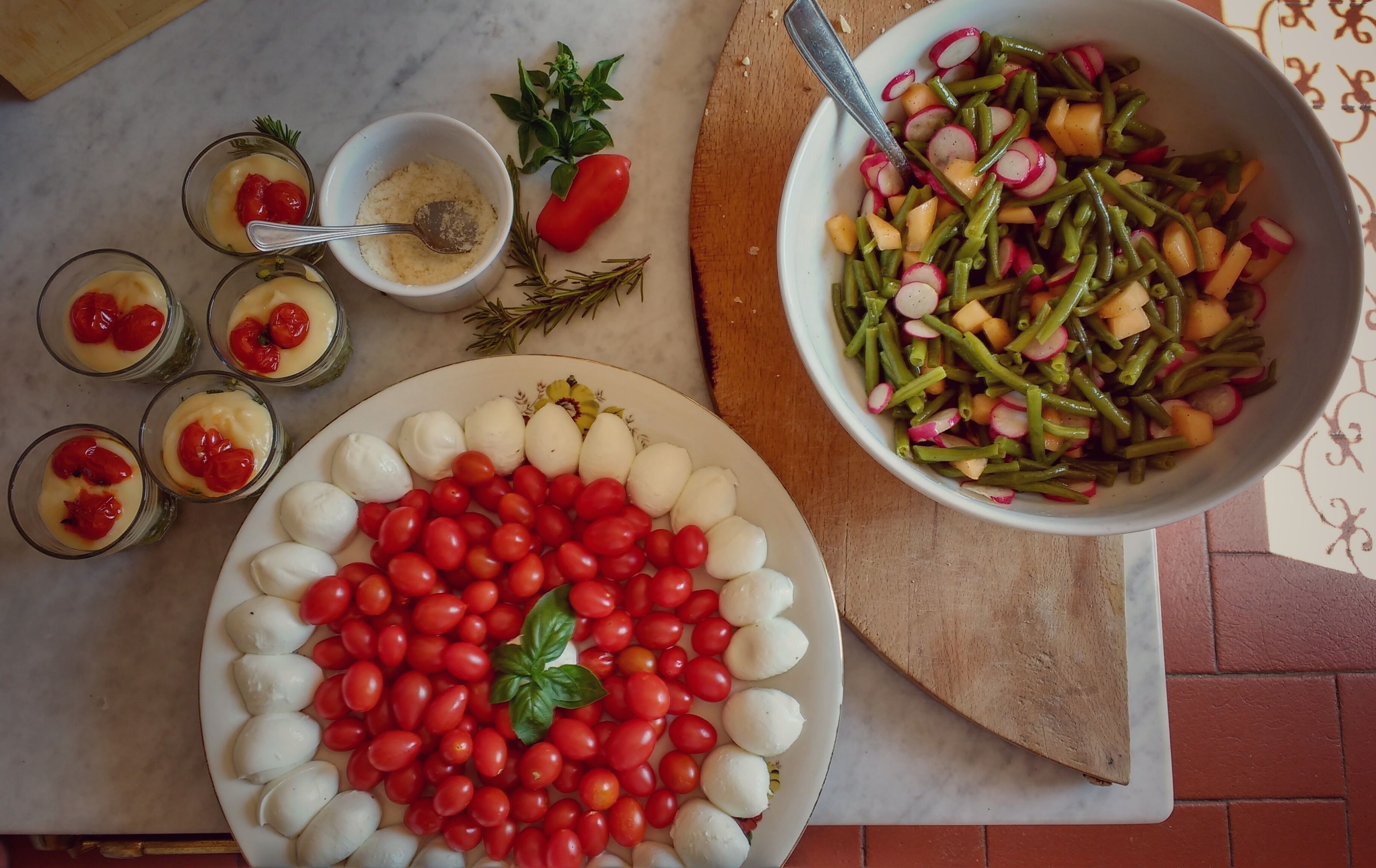 A table with a plate of mozzarella balls and cherry tomatoes, a bowl of green beans and radishes, and small glasses of a creamy dessert with cherries.