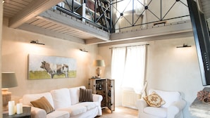 Ein geräumiges Wohnzimmer mit hoher Decke, einem weißen Sofa, einem kleineren weißen Sessel, einem Couchtisch und einem Gemälde einer Kuh.