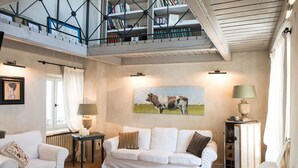 Ein geräumiges Wohnzimmer mit hoher Decke, einem großen Gemälde von einer Kuh und einer Treppe, die zu einem mit Büchern gefüllten Dachboden führt.