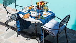 Ein Tisch, gedeckt für eine Mahlzeit am Pool, mit einer tischgetüchelten Tischdecke in Punkten, Metallstühlen und einer Auswahl an Speisen und Getränken.