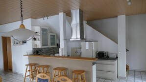 Una cocina moderna con una isla central, taburetes de bar, una lámpara colgante y una escalera que sube a otro nivel.