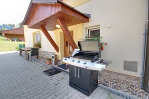 Una parrilla para barbacoa al aire libre con una pérgola de madera encima, una planta en maceta y una pequeña mesa con sillas.