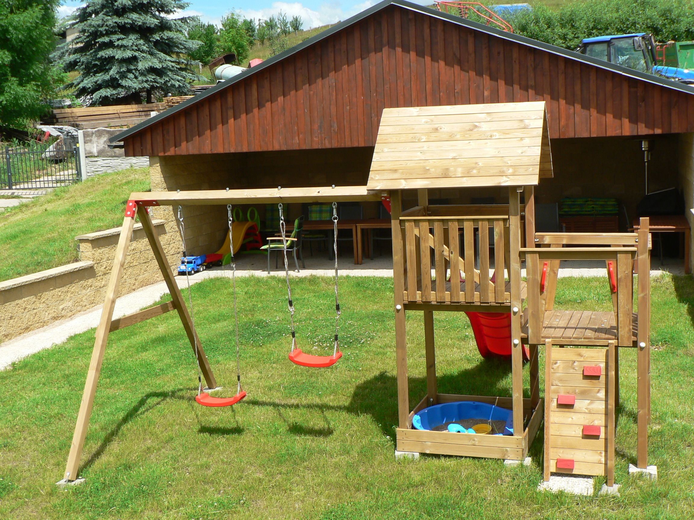 A wooden play structure with swings and a slide, set against a building with a sloped roof.