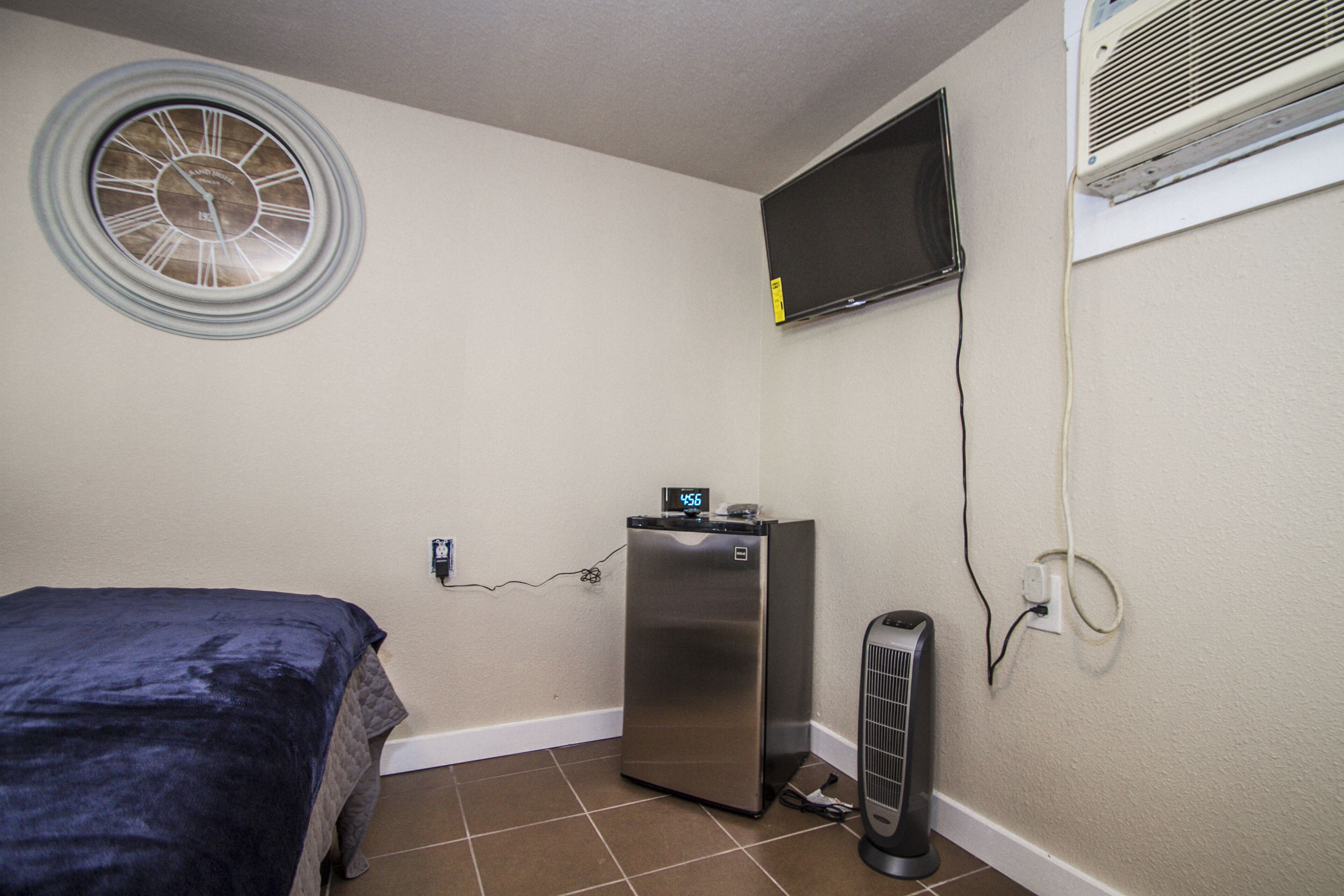 Ein kleines Zimmer mit Bett, Wanduhr, Fernseher, Kühlschrank und Heizung.
