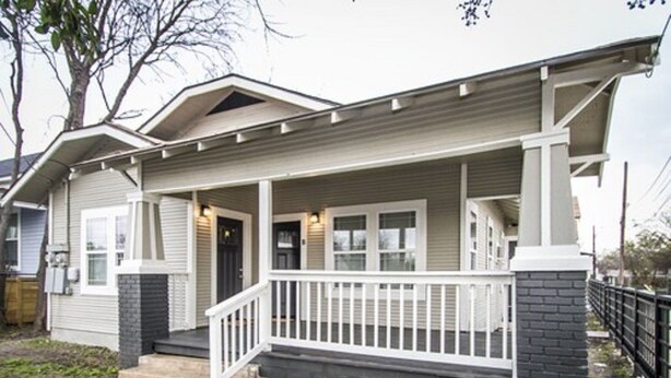 Una casa de dos pisos con un porche delantero, base de ladrillo y un pequeño jardín frontal.