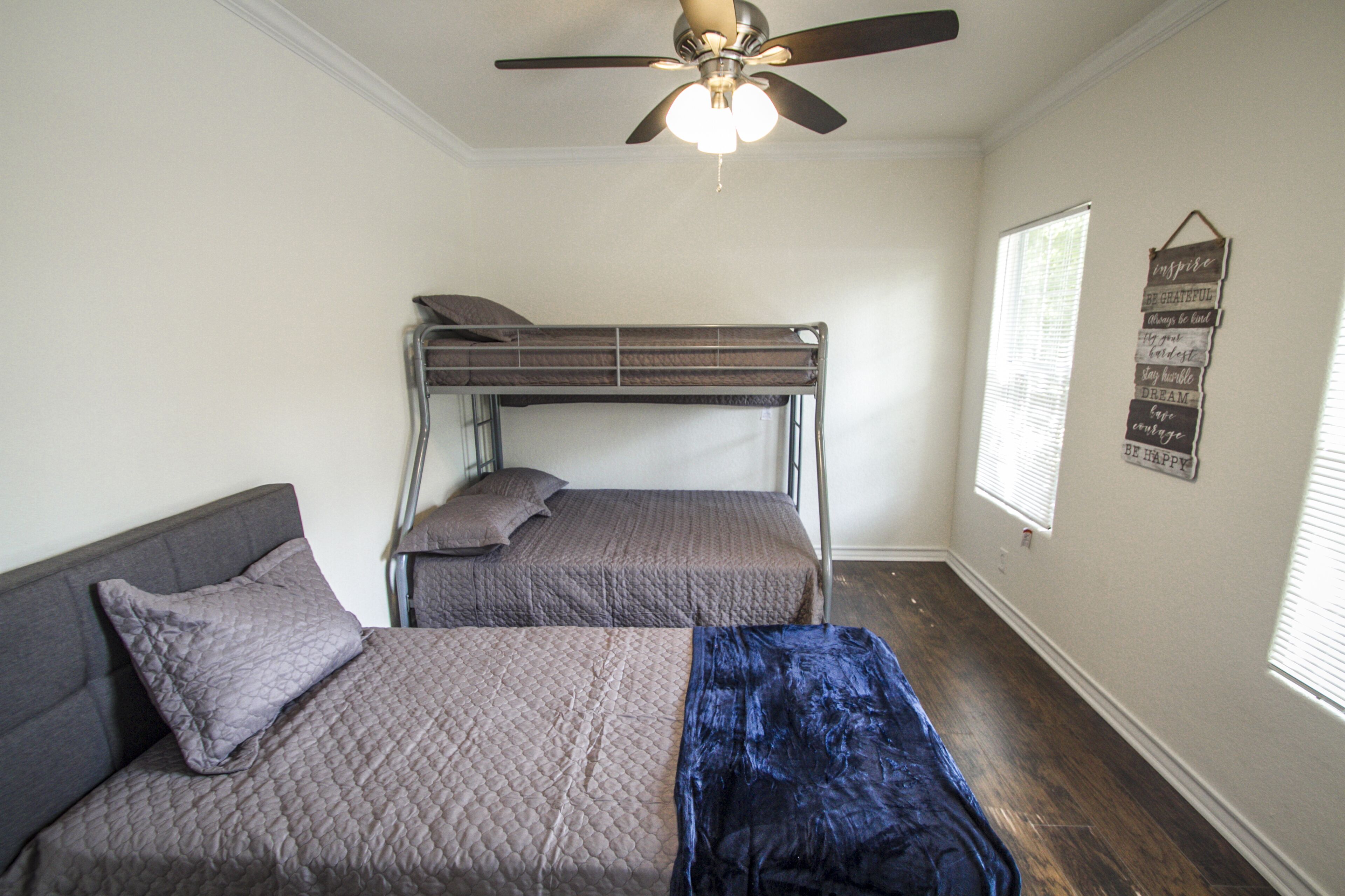 A bedroom with a bunk bed, a single bed, a ceiling fan, and a motivational quote on the wall.