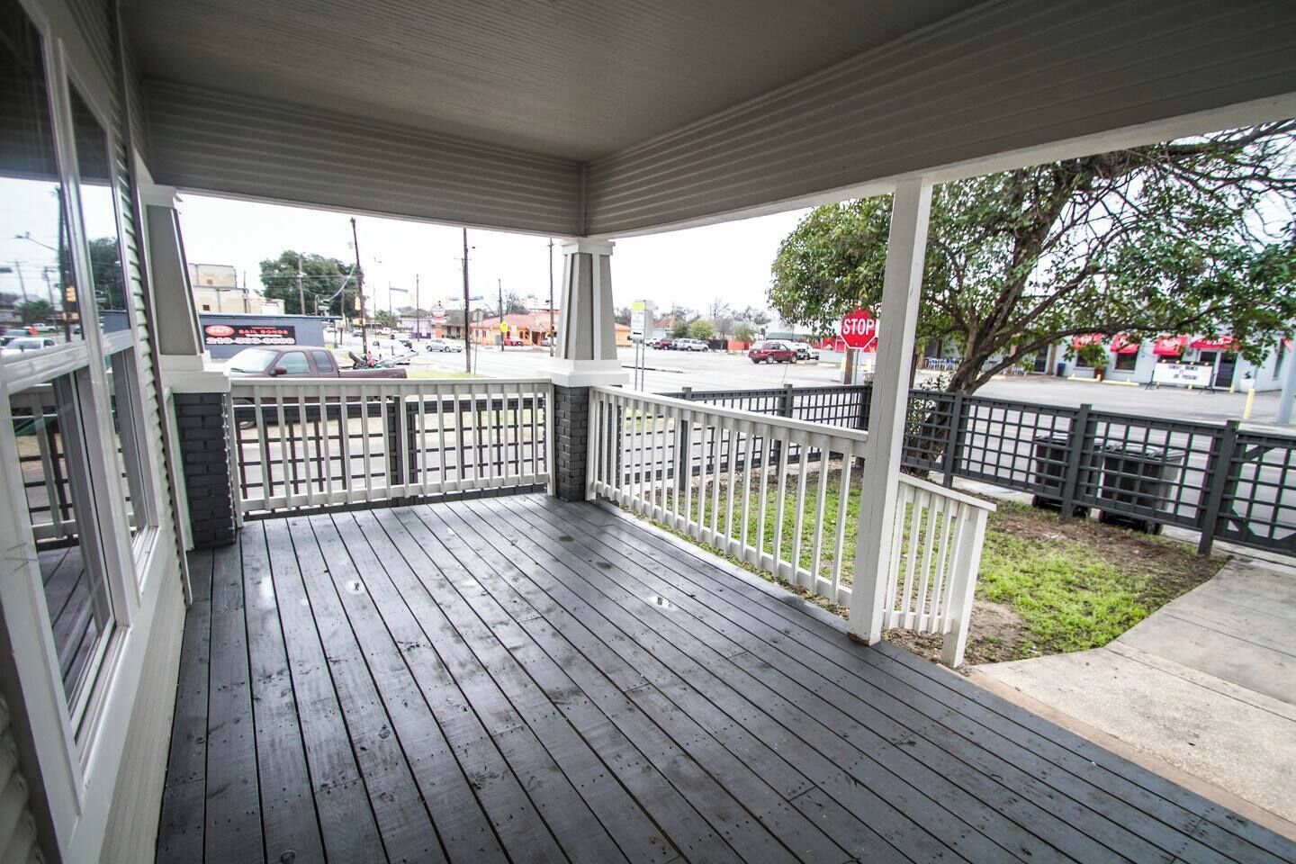A covered porch with a wooden deck and white railings, overlooking a street with parked cars and a stop sign.