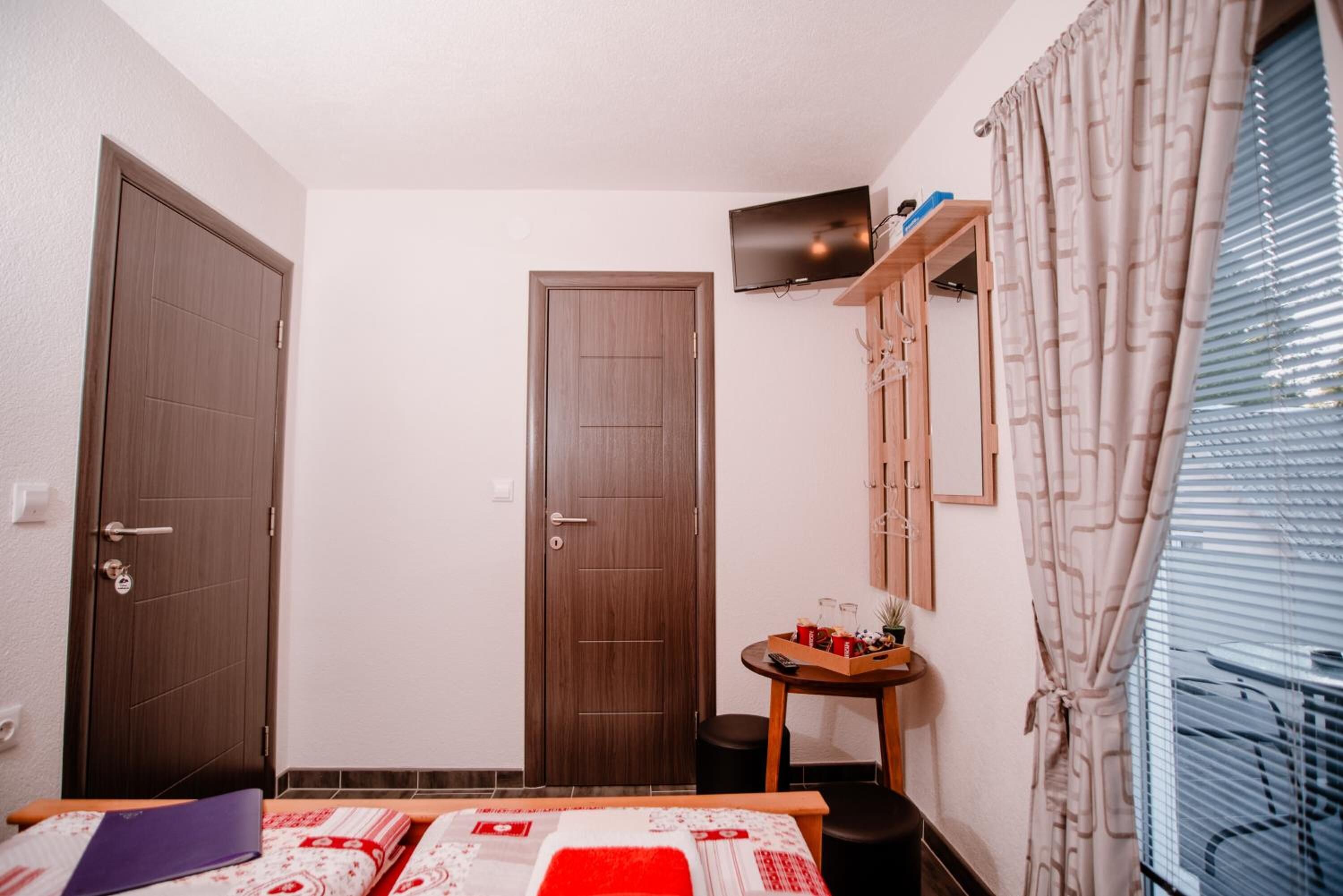 Una habitación pequeña y ordenada con dos puertas, una cama con colcha roja y blanca, una mesita con una caja de aperitivos, un televisor en un estante y una ventana con persianas.