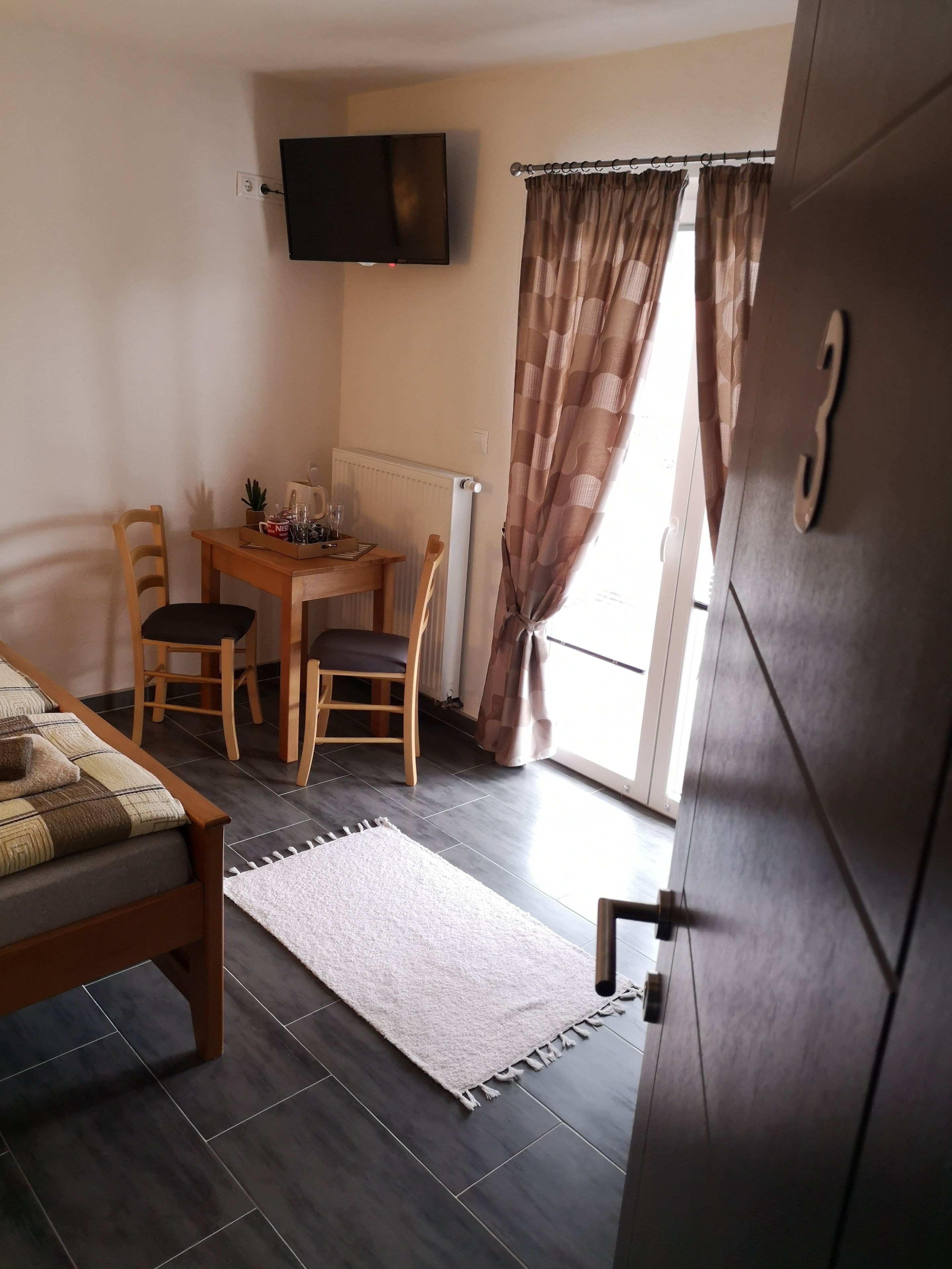Una habitación pequeña y bien iluminada, con una cama, un escritorio y una silla. Hay un televisor de pantalla plana montado en la pared, un radiador debajo del escritorio y una ventana con cortinas.