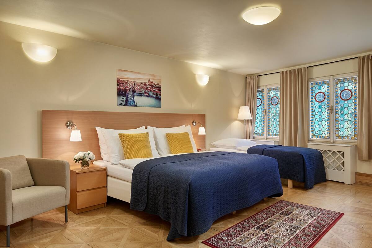 A hotel room with two beds, a nightstand, a chair, and a large window with stained glass.
