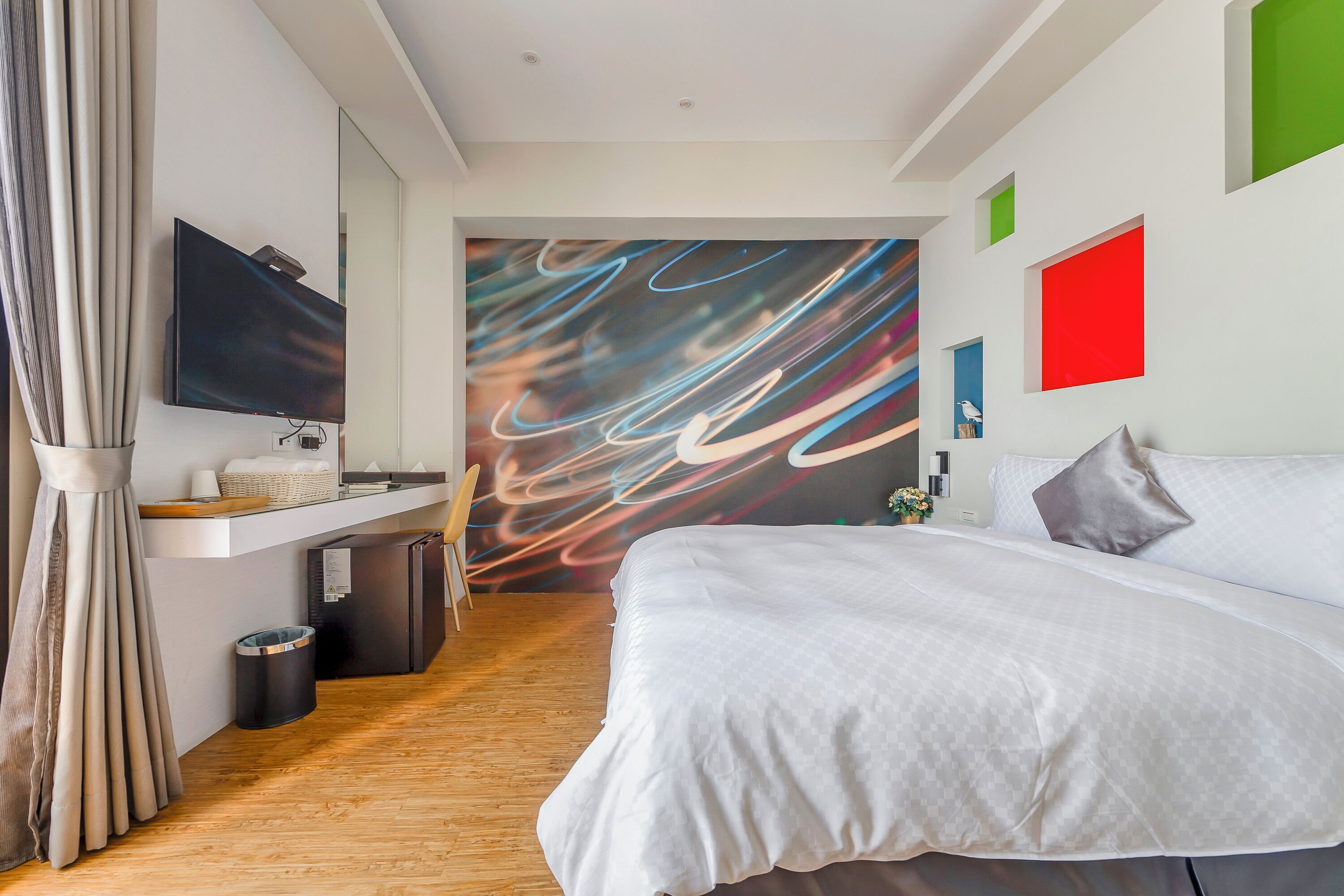 Een moderne hotelkamer met een groot bed, een flatscreen televisie aan de muur, een klein bureau met stoel, en een kleurrijke abstracte muurschildering.