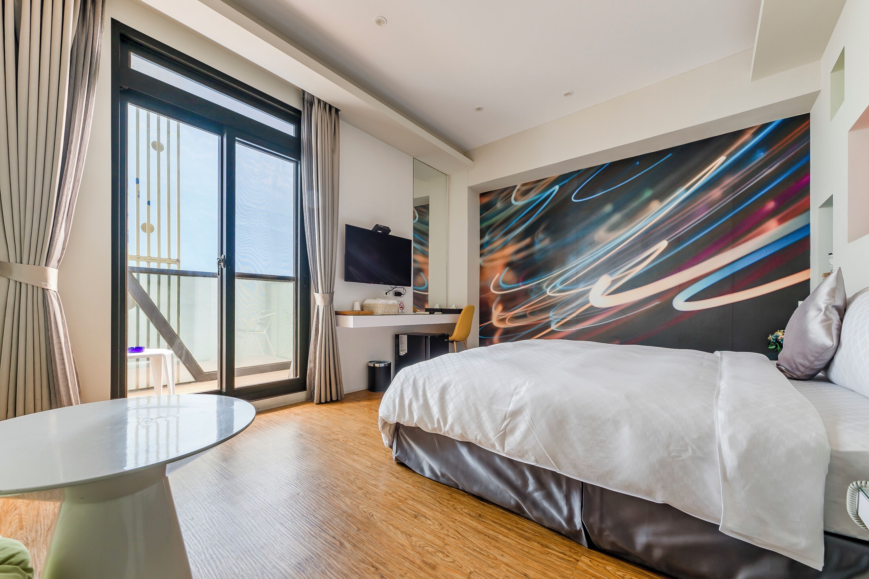 A modern hotel room with a large bed, a desk, a TV, and a balcony with a view.