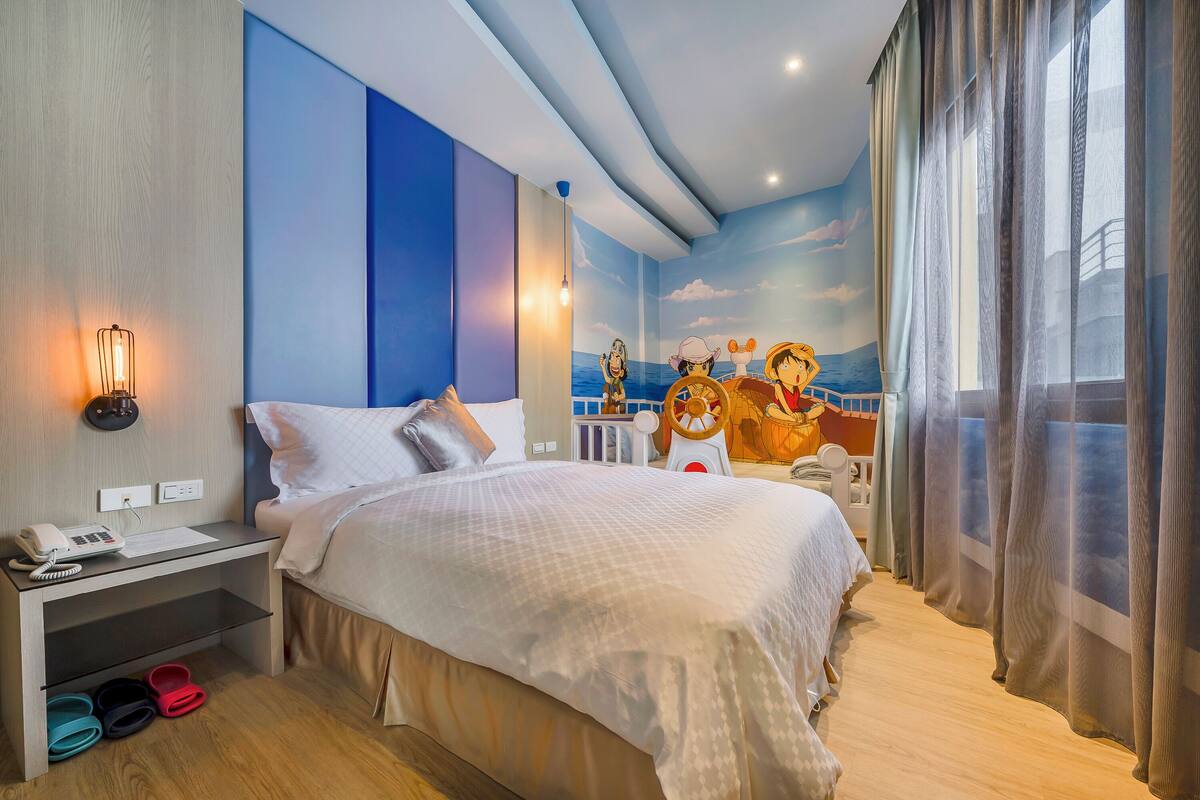 A modern hotel room with a large bed, a nightstand with a phone, slippers, and a mural of a ship on the wall.