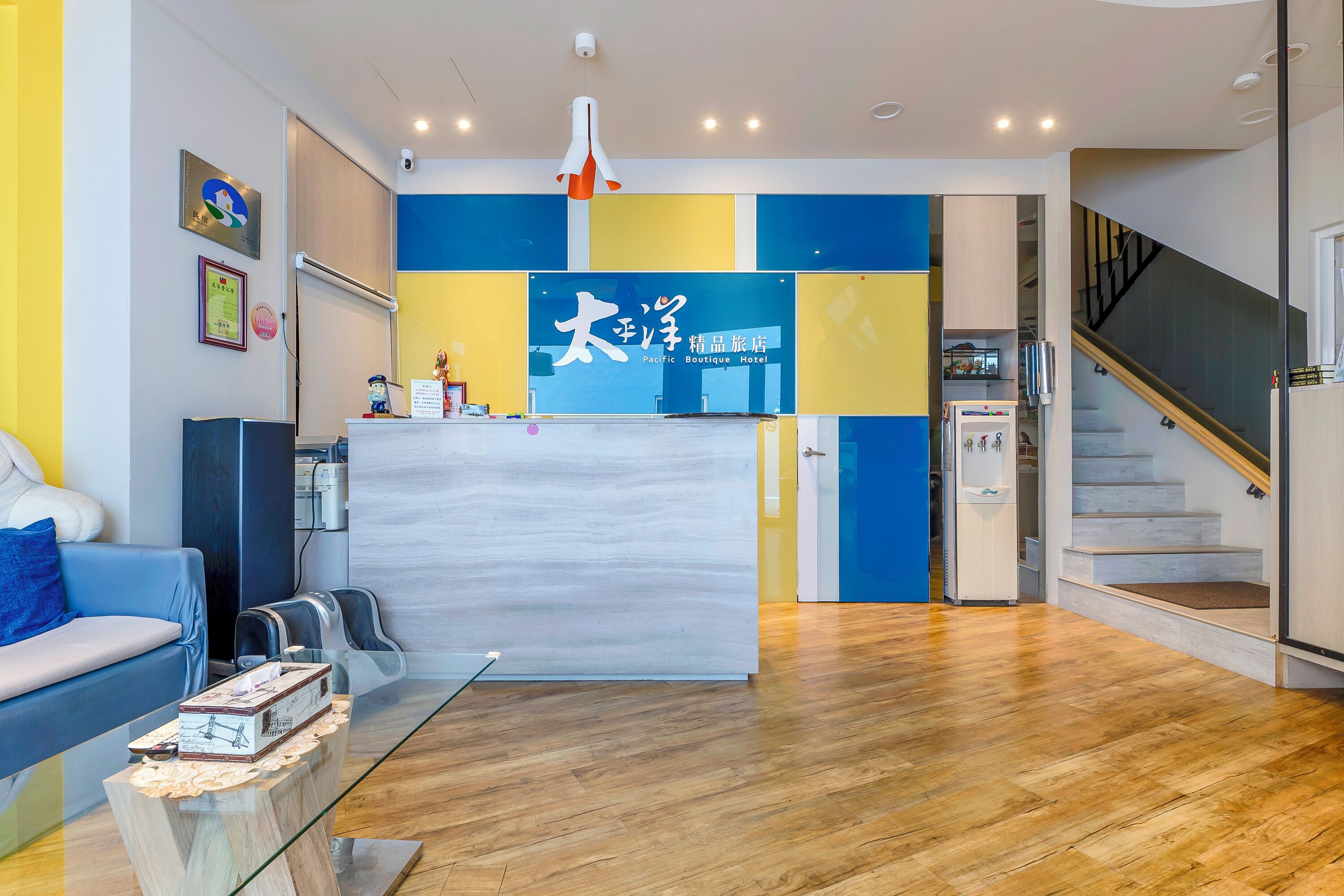 A hotel reception area with a modern design, featuring a reception desk, a blue and yellow color scheme, a staircase, and a wooden floor.