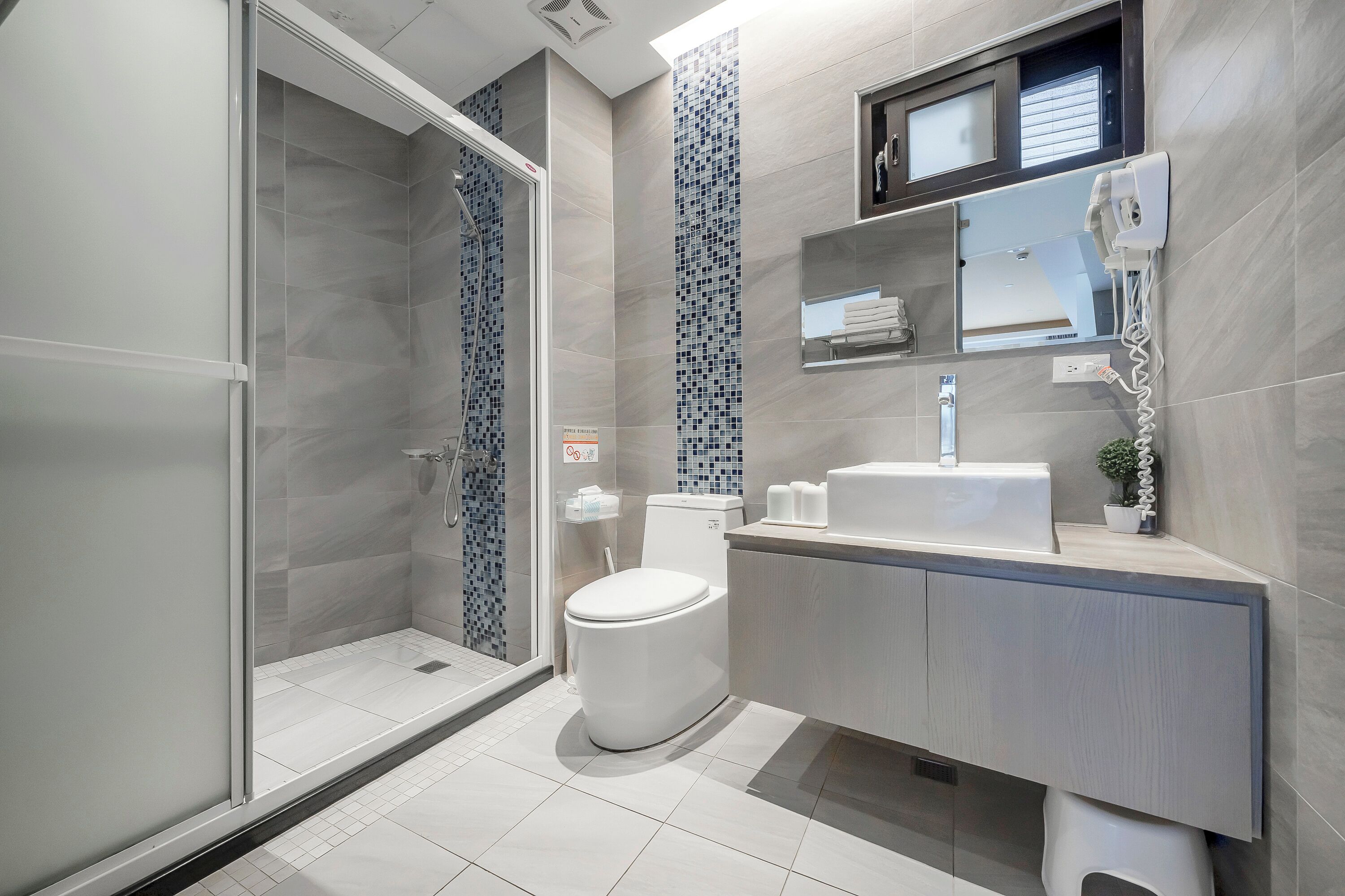 A modern bathroom with a glass shower enclosure, a toilet, a sink with a mirror above, and a small shelf with toiletries.