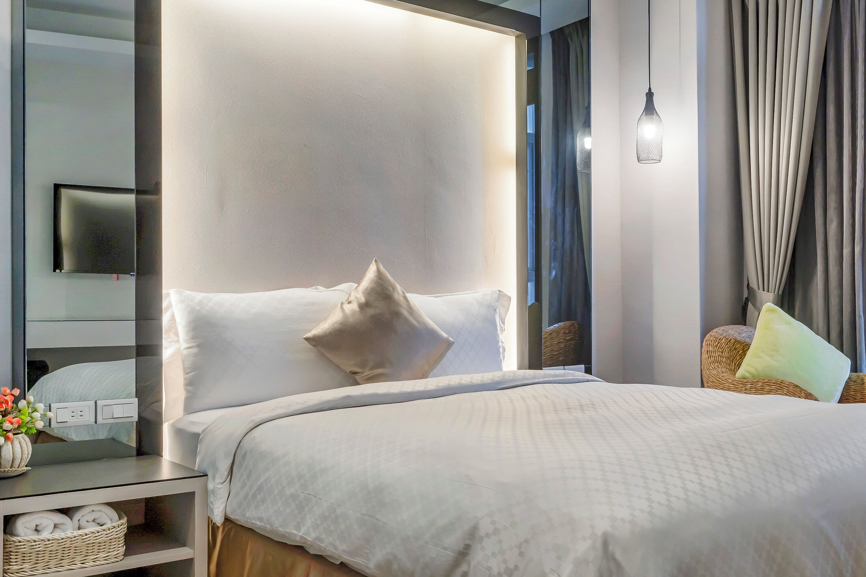 Une chambre d’hôtel moderne avec un lit soigneusement fait, une table de chevet ornée d’un vase de fleurs et un téléviseur à écran plat fixé au mur.