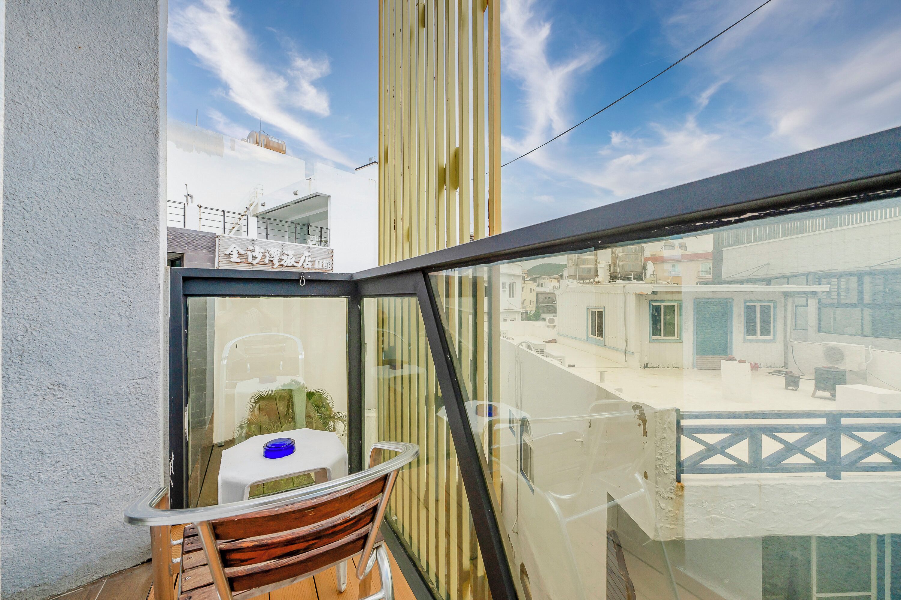 A balcony with a wooden chair and a small table, a glass railing, and a view of buildings and a bridge in the background.