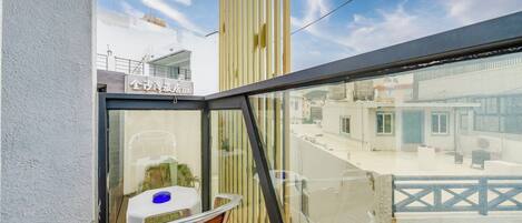 A balcony with a wooden chair and a small table, a glass railing, and a view of buildings and a bridge in the background.