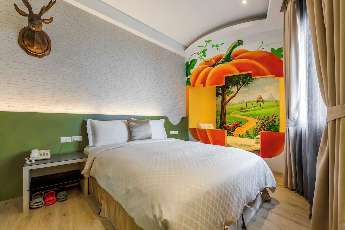 A neatly made bed with a white comforter, a nightstand with a phone and slippers, a wall-mounted deer head decoration, and a mural of a pumpkin field.