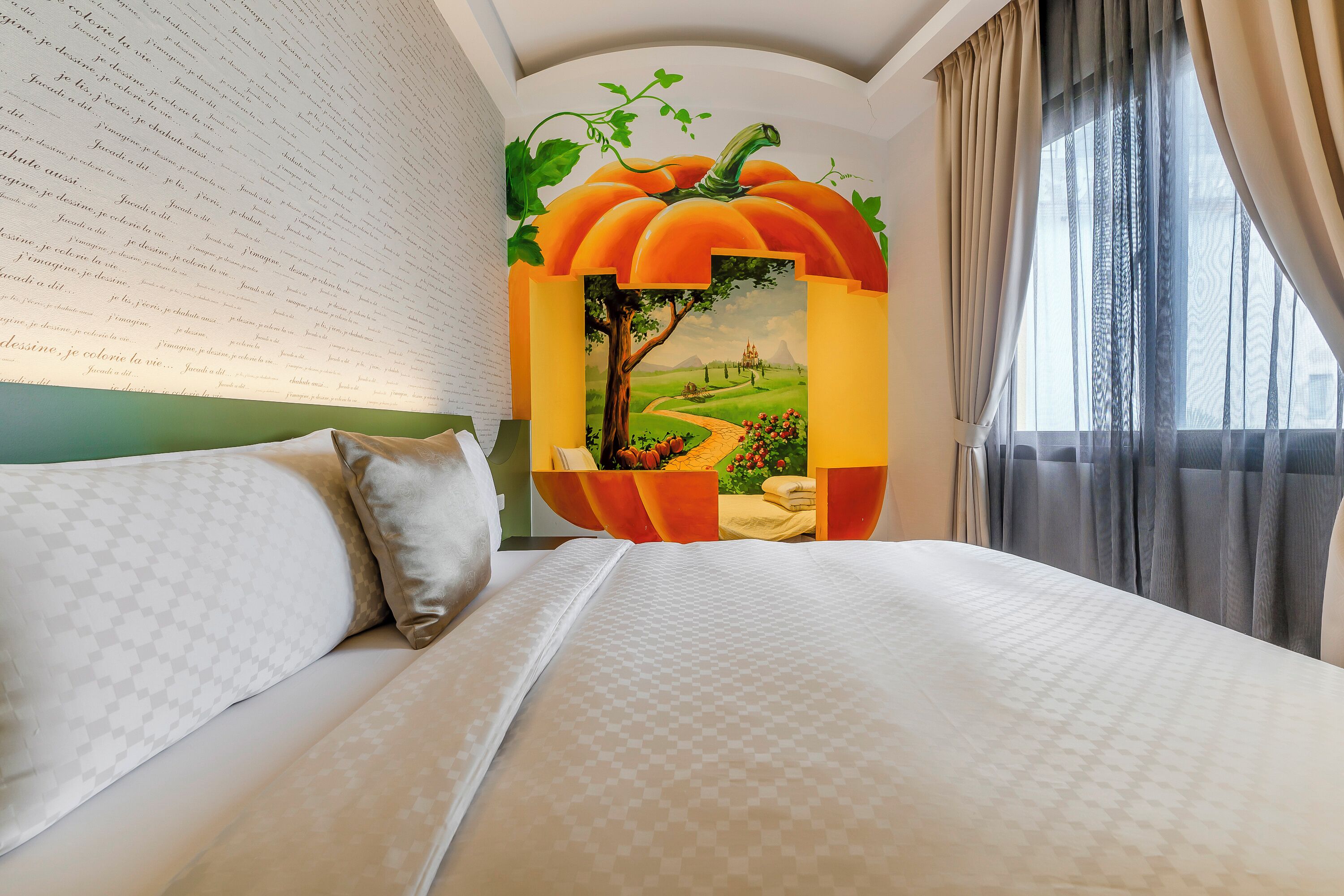 A neatly made bed with a patterned bedspread, a wall with a mural of a pumpkin and a landscape, and a window with sheer curtains.