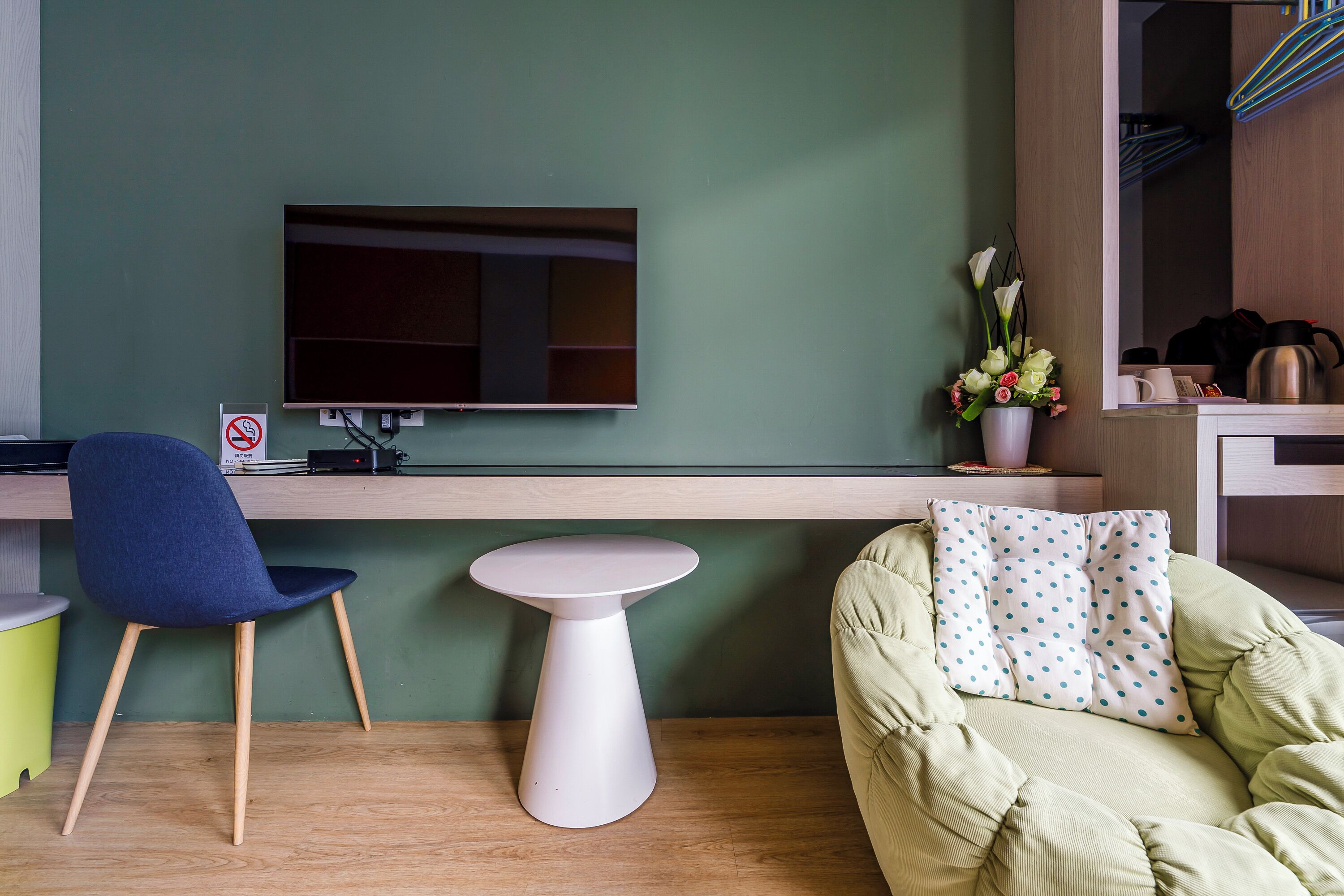 A modern room with a green wall, a flat-screen TV mounted above a wooden desk, a blue chair, a white round table, and a green sofa with a polka dot pillow.