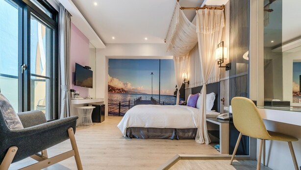 A modern hotel room with a large bed, a desk, a chair, and a view of the sea through a large window. - Pacific Inn (Hengchun)