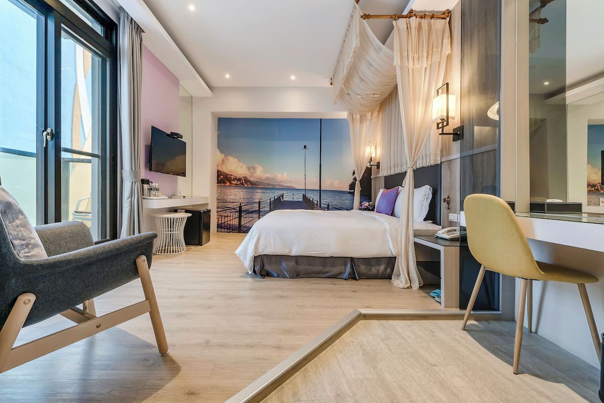 A modern hotel room with a large bed, a desk, a chair, and a view of the sea through a large window.