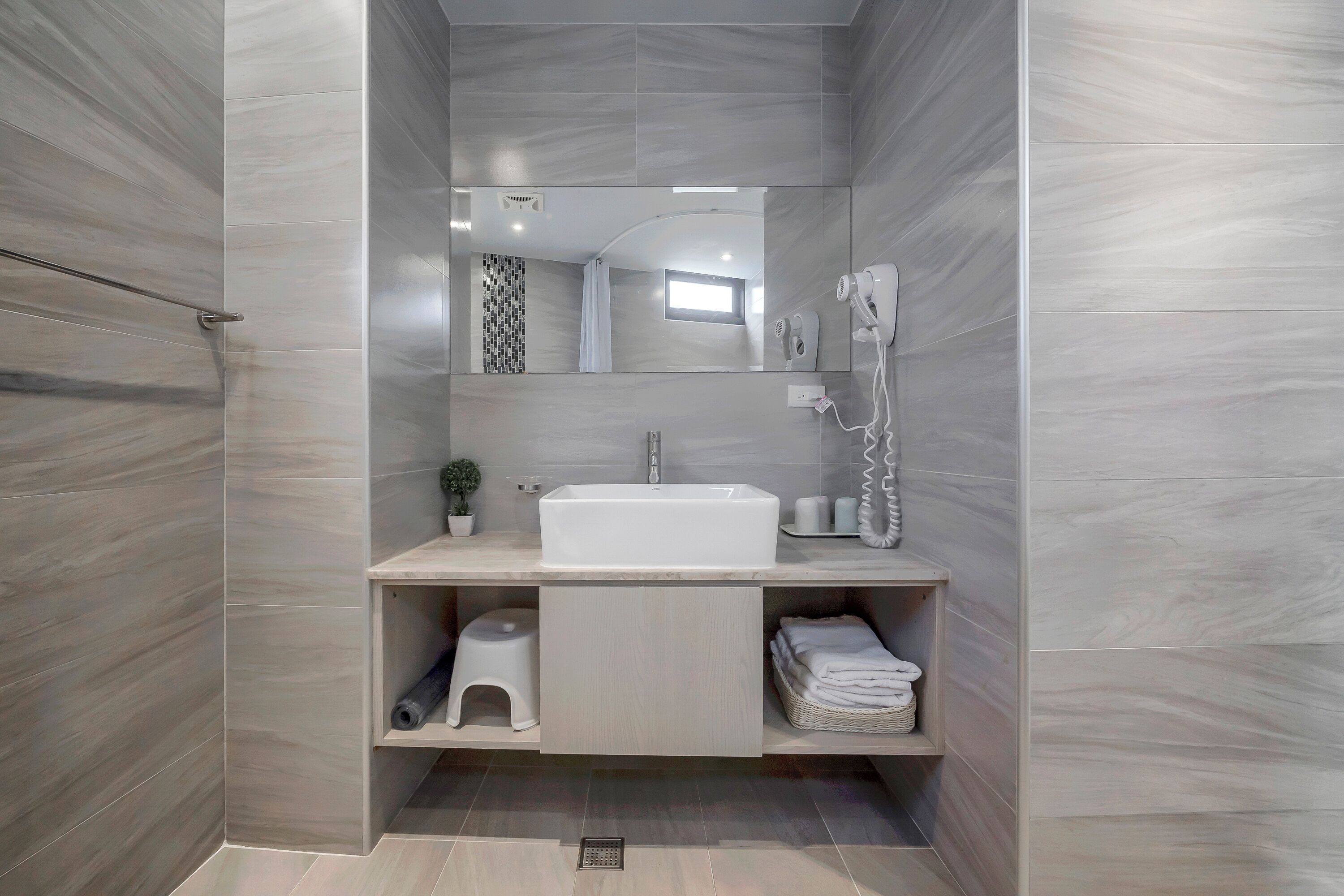 A modern bathroom with a large mirror, a sink, and a shower. There are shelves with towels and toiletries.