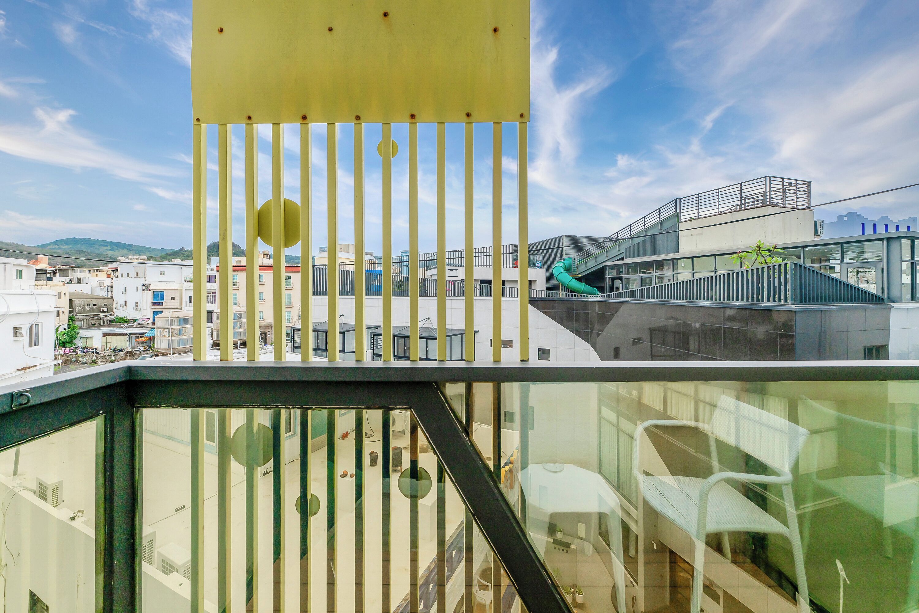 A modern building with a glass facade, a yellow structure with vertical bars, and a view of a cityscape with buildings and a green hill in the background.