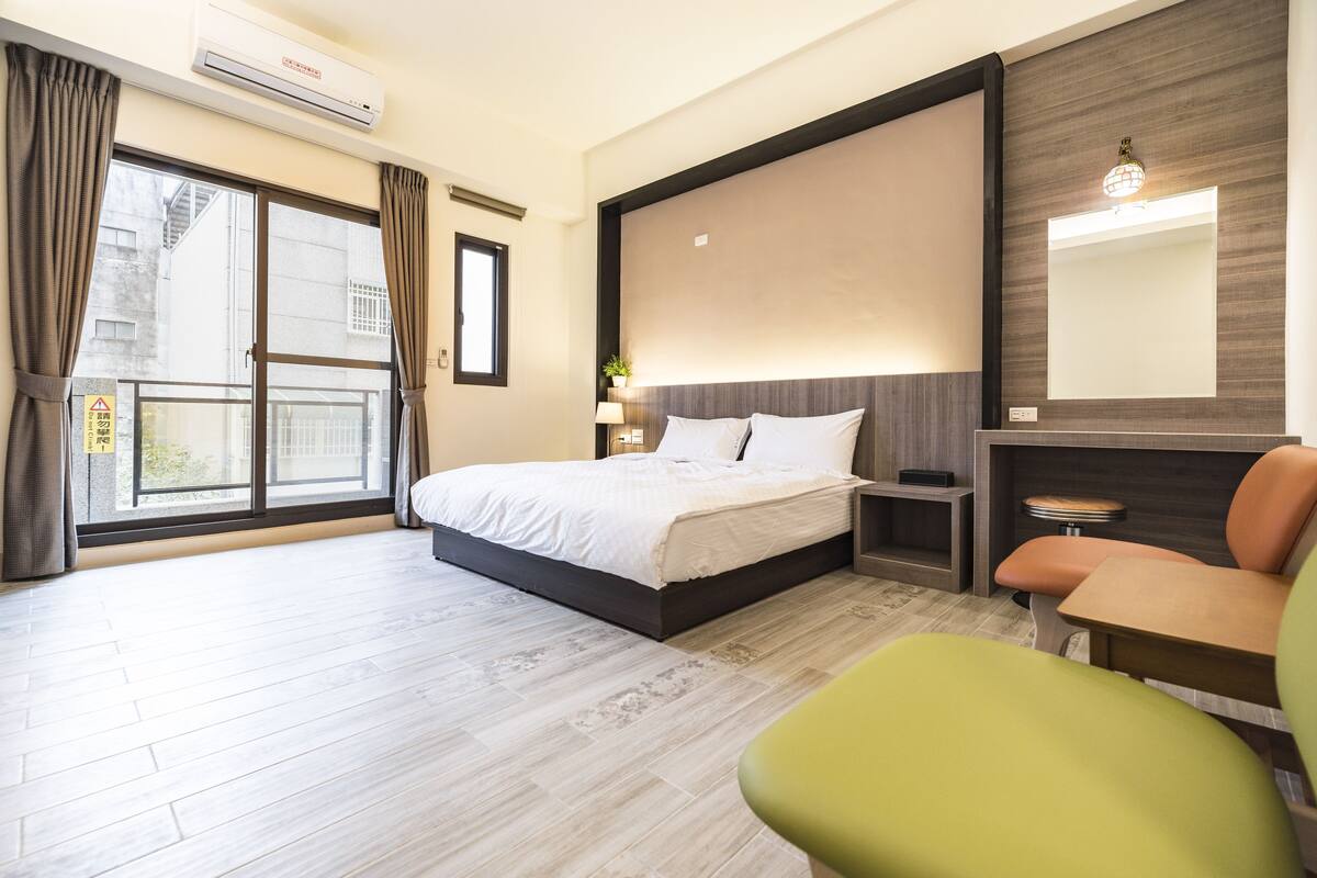 A hotel room with a bed, a desk, a chair, a large window, and a wall-mounted air conditioner.